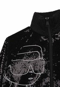 Giacca nera con zip e colletto a coste, impreziosita da paillettes, con un design grafico bianco che rappresenta un volto con occhiali da sole.