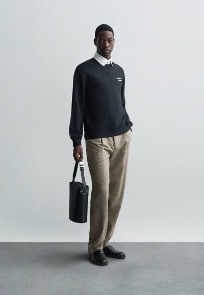 Čierny sveter s bielym logom, biela košela s golierom pod ním, hnedé vzorované nohavice a čierna cylindrická taška. Čierne kožené topánky.