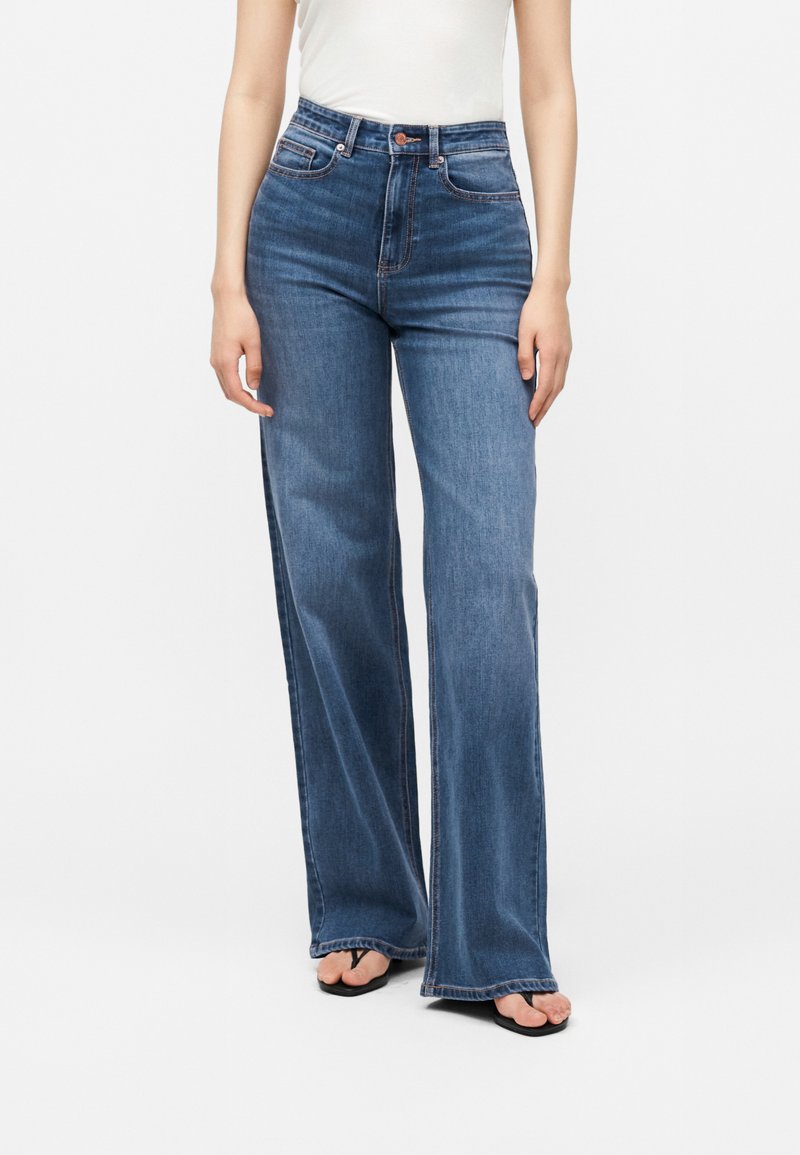 Femme portant un jean bleu taille haute à jambes larges et des tongs noires, debout devant un fond blanc, les bras détendus le long du corps.