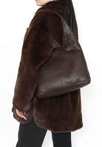 Borsa hobo in pelle marrone con una texture morbida, forma rotonda e una sola tracolla, indossata sopra un cappotto in pelliccia sintetica marrone scuro.