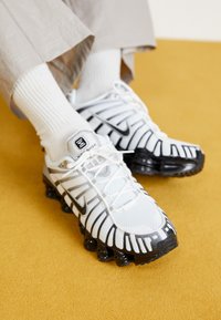 Hvit og svart atletiske sko med mesh-overdel, gummisåle og distinkte ribbestrikkede detaljer. Brukt med hvite ribbestrikkede sokker.