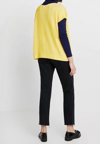 Žlutý žebrovaný pletený top nošený přes tmavě modrou košili s dlouhým rukávem, kombinovaný s černými zkrácenými džínami a černými botami, pohled zezadu.