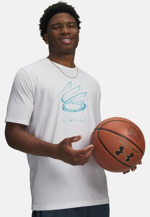 Jonge man die lacht, een witte Curry T-shirt en een gouden ketting draagt, met beide handen een Under Armour basketbal vasthoudt.