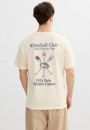 Hombre con camiseta beige con el diseño "Whitehall Club United Kingdom Team 1924 Paris Olympic Games" y remos cruzados en la parte trasera.