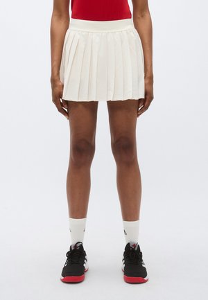 Falda crema plisada con cintura elástica, combinada con zapatillas deportivas negras y calcetines blancos con detalles negros.