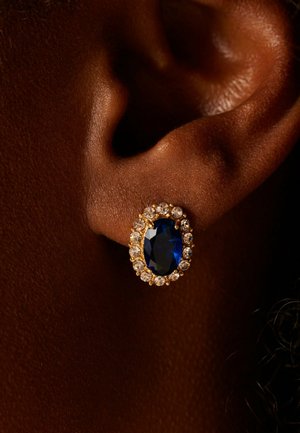 Gouden oorbel met ovale diepblauwe edelsteen omringd door kleine heldere kristallen, gedragen aan een donkergekleurde oor.