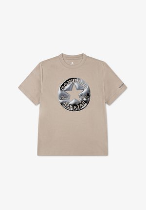 T-shirt beige à manches courtes avec un grand logo Converse All Star argenté métallique et le texte "Chuck Taylor" sur le devant.