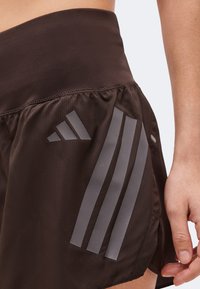 Κοντινό πλάνο ενός ατόμου που φοράει σκούρα καφέ αθλητικά σορτς με αντανακλαστικές τρεις ρίγες και λογότυπο στο πλάι, το χέρι ακουμπά κοντά στο τελείωμα.