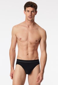 Slip nero con elastico a contrasto, caratterizzato da una texture liscia e un design aderente. Modello con un fisico muscoloso.