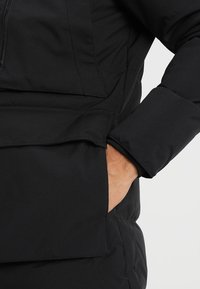 Černá izolovaná bunda s kapsou na zip a bočním rozparkem pro přístup rukou, vyrobená z hladké, odolné látky.