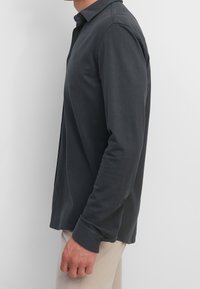 Langarm Polo-Shirt in Dunkelgrau, aus Baumwolle gefertigt. Mit einem Kragen, Knopfverschluss und seitlichen Belüftungen. Glatte Textur und figurbetonter Schnitt.