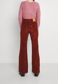 Pantalon évasé en velours côtelé rouge avec poches arrière et une étiquette sur la ceinture. Le modèle porte un pull rose texturé.