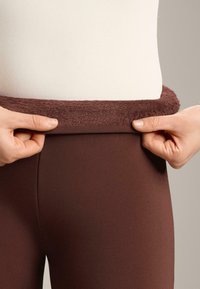 Hnedé legíny s hrubým, jemne chlpatým pásom; ruky upravujú pás. Materiál vyzerá hladko a pružne.