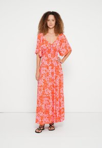 Gara, plūstoša kleita spilgti oranžā krāsā ar rozā ziedu rakstiem. Kleitai ir īsas, pūkainas piedurknes un savākts korsete. Valda ar melnām sandales ar siksniņām.