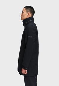 Giacca nera impermeabile con colletto alto, chiusura a zip e tasche laterali. Presenta un logo sulla manica e un design elegante e minimalista.