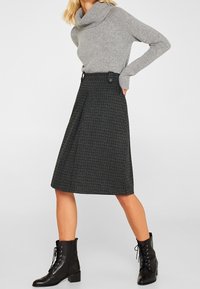 Jupe à carreaux noir et gris avec des plis, taille haute et boutons ; associée à un pull gris clair et des bottines noires.