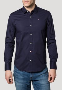 Navy button-up shirt made of smooth fabric, featuring a classic collar, long sleeves, and a small logo on the chest. White buttons.