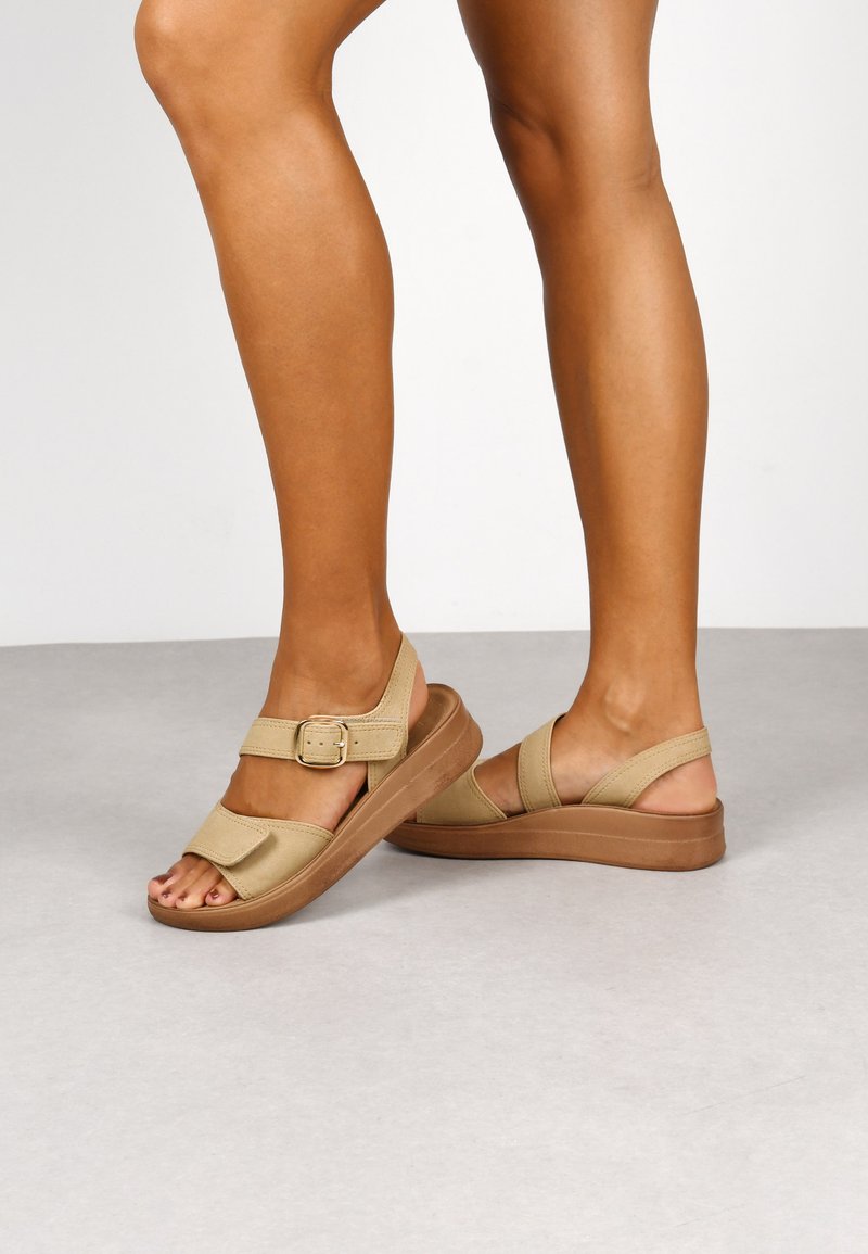 Sandalias beige con un acabado texturizado, con una correa ancha y hebilla ajustable, sobre una suela de plataforma marrón claro.