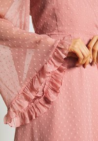 Mains de femme jointes près de la taille, portant une robe texturée rose avec des manches cloche transparentes et volantées ainsi que de petits motifs carrés.