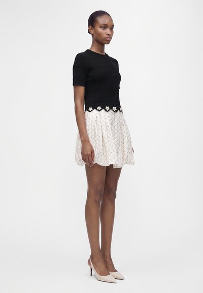 Jeune femme debout portant un haut noir à manches courtes avec bordure florale et une jupe courte blanche à motifs avec des talons beiges.