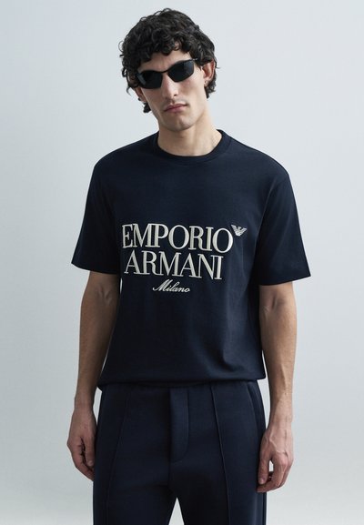 Marineblauw katoenen T-shirt met het logo "Emporio Armani Milano" in lichtcrème. Het model draagt ook een zwarte zonnebril. Eenvoudig ontwerp met een comfortabele, losse pasvorm.