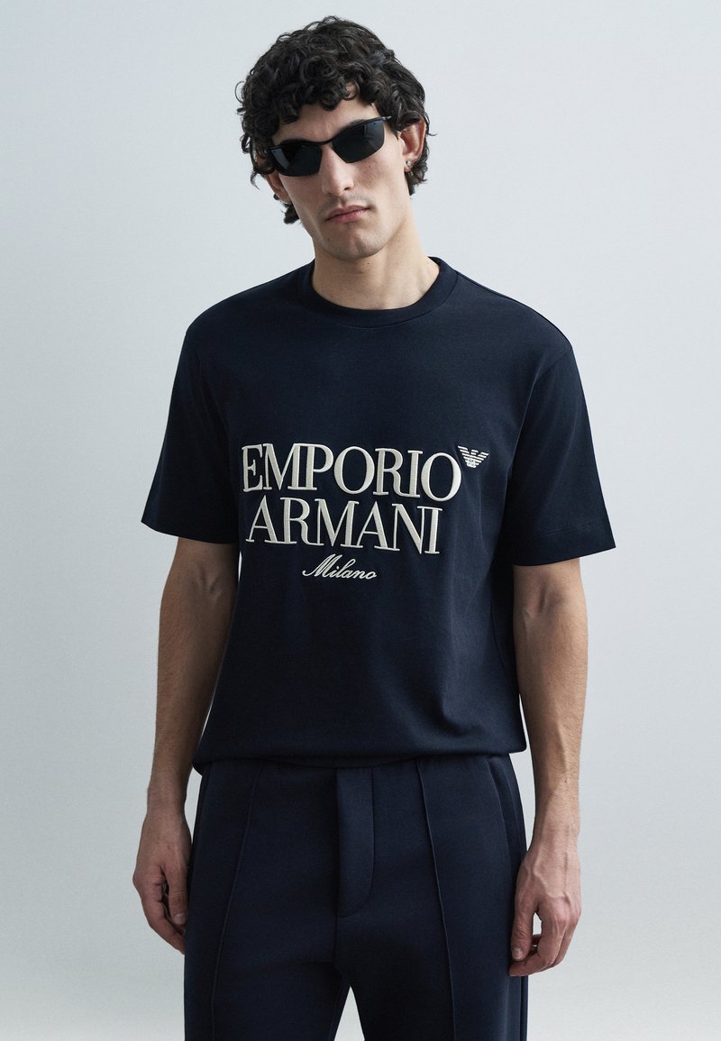Μπλούζα από βαμβάκι σε ναυτικό μπλε χρώμα με το λ�ογότυπο «Emporio Armani Milano» σε ανοιχτό κρεμ χρώμα. Το μοντέλο φορά επίσης μαύρα γυαλιά ηλίου. Απλός, χαλαρός σχεδιασμός εφαρμογής.