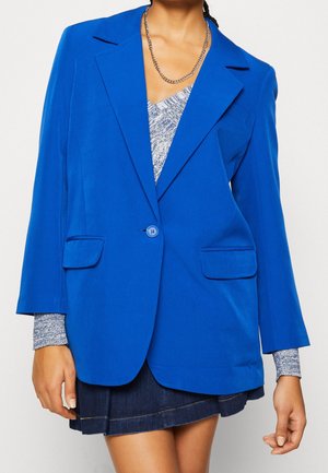 Personne portant un blazer bleu vif sur un haut gris à motifs, associé à une jupe en jean foncé et un collier chaîne en argent.