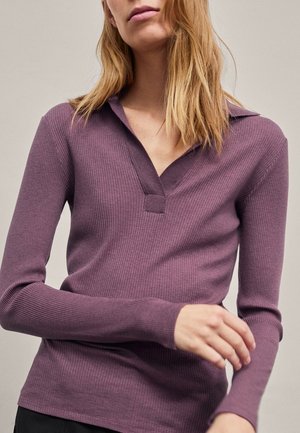 Vrouw draagt een paarse, geribbelde, lange mouw trui met V-hals en past de mouw aan tegen een effen achtergrond.