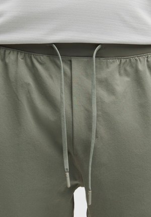 Shorts atletici verdi da uomo con vita elastica e cordino. Il tessuto è morbido, con una consistenza liscia e presenta tasche laterali.
