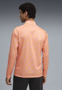 Dlhý rukáv košele z ružovej látky s geometrickým žltým vzorom. Dizajn obsahuje vysoký golier a hladkú, ľahkú textúru.