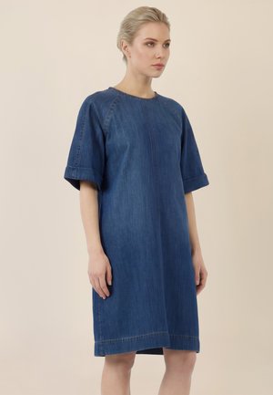 Frau mit hellen, zurückgebundenen Haaren, die ein locker sitzendes, knielanges blaues Denimkleid mit ellbogenlangen Ärmeln trägt, steht vor einem beigen Hintergrund.