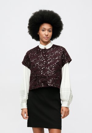 Ženska z afro pričesko nosi belo bluzo z dolgimi rokavi, kratko bleščečo temno vijolično jakno z kratkimi rokavi in črno krilo na belem ozadju.