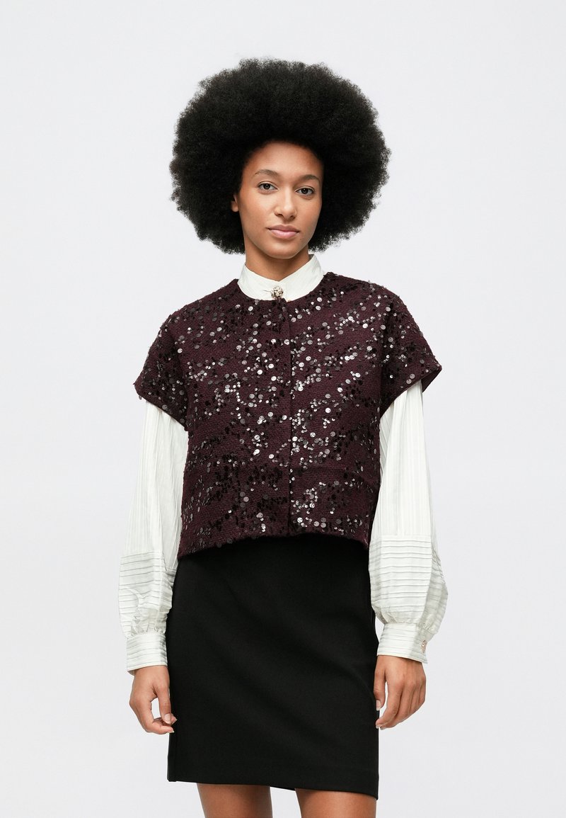 Ženska z afro pričesko nosi belo bluzo z dolgimi rokavi, kratko bleščečo temno vijolično jakno z kratkimi rokavi in črno krilo na belem ozadju.