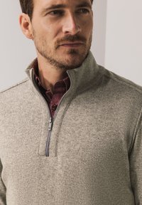 Grauer Herrenstrickpullover mit Reißverschluss aus weichem Strickstoff, ausgestattet mit einem hohen Kragen und einem darunter sichtbaren bordeauxfarbenen Hemd mit Knöpfen.