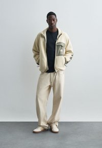 Chaqueta de forro polar en color crema con un bolsillo en el pecho verde y logo, combinada con pantalones beiges y zapatos de color claro con rayas.