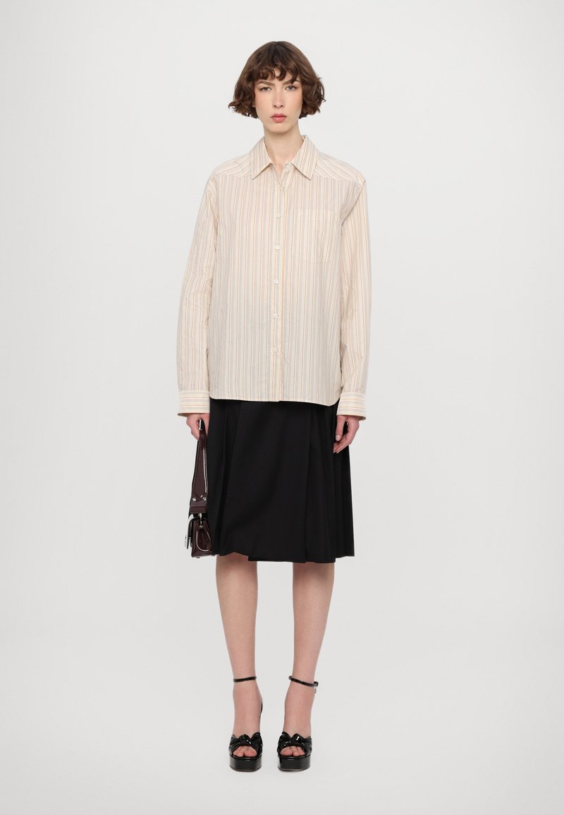 Camisa de botones a rayas en colores claros, combinada con una falda plisada negra. La modelo sostiene un bolso oscuro y lleva sandalias de plataforma negras.