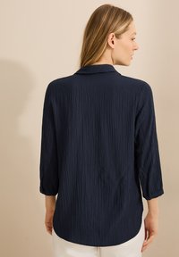 Donkerblauwe shirt met een gestructureerde, gekreukte stof. Heeft een kraag en driekwartmouwen, met een ronde zoom aan de achterkant.
