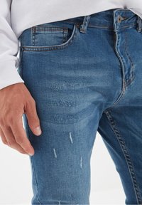 Jean bleu délavé avec des détails usés, présentant une coupe slim, un design à cinq poches et une fermeture par bouton à la taille.