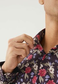 Korte mouwen bloes met een donkerpaarse stof versierd met rode en witte bloemen, groene bladeren en grijze knopen op de kraag.