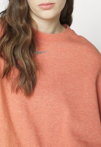 Woman wearing a coral sweatshirt with a small glittery Nike logo near the collar, showing long wavy brown hair and lips.