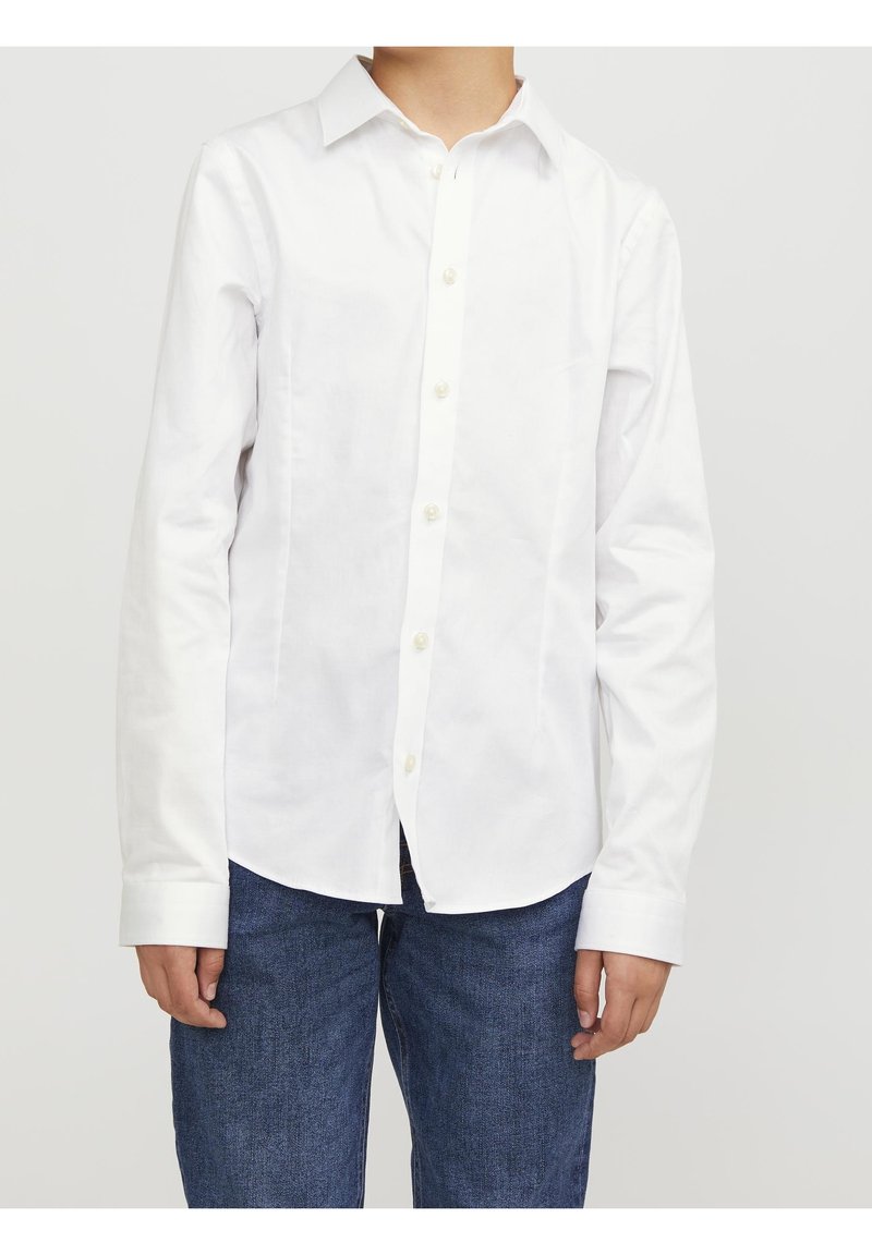 Personne portant une chemise blanche à manches longues boutonnée et un jean bleu, debout devant un fond clair uni.