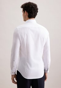 Uomo con capelli ricci scuri che indossa una camicia bianca attillata e pantaloni scuri, in piedi di spalle contro uno sfondo chiaro e semplice.