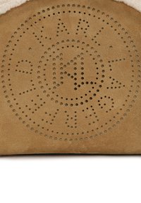 Tan semsket stoff har et preget sirkulært logo og perforert design over overflaten, med en myk beige fôr øverst.