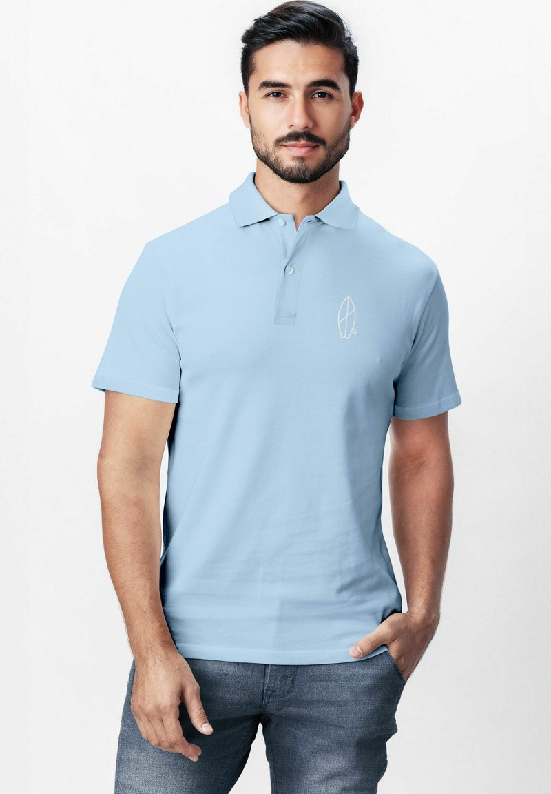 Helles blaues Poloshirt aus weichem Stoff, mit klassischem Kragen und kleinem weißen Logo auf der linken Brust, kombiniert mit grauen Jeans.