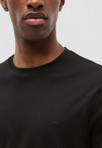 Primer plano de una persona que lleva una camiseta negra de cuello redondo con un pequeño logo bordado de "MK" en el pecho.