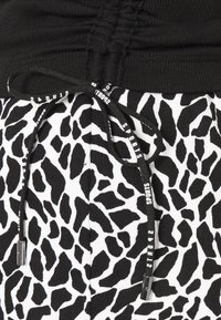 Gros plan sur un pantalon imprimé animal noir et blanc avec une ceinture élastique noire et des cordons noirs portant l’inscription « SPORTS ».