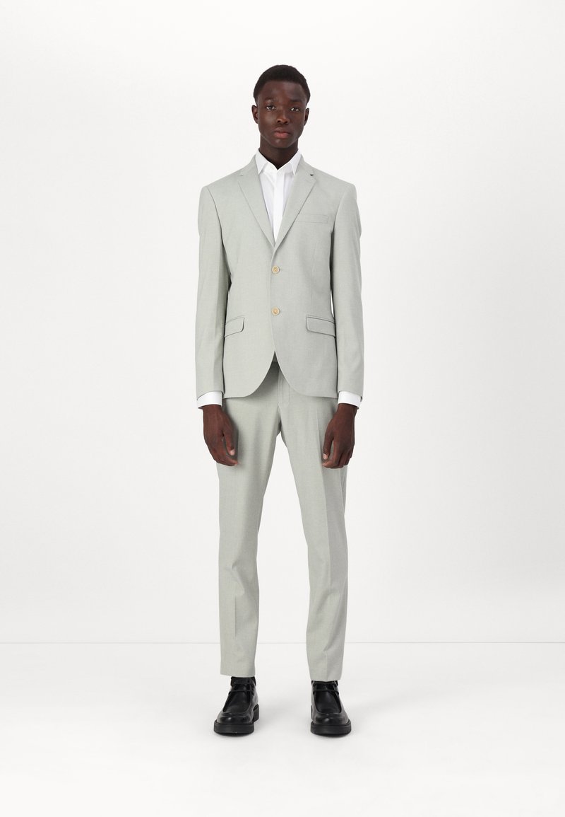 Šviesiai pilkas kostiumas, pritaikytas tinkamai, su dviem mygtukais ir išpjauta apykakle. Derinamas su balta marškiniais ir juodais batais. Lygus paviršius.