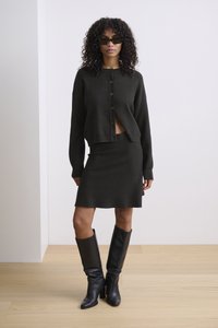 Tamnozelena pletena kardigan sa dugmadima, krojen najprije, uparen sa odgovarajućom uskom suknjom. Model nosi čizme do koljena u crnoj boji.