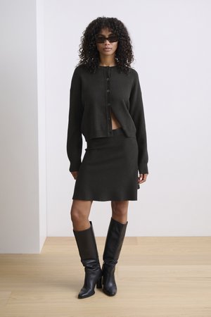 Dark green knit cardigan with buttons, cropped design, paired with a matching fitted skirt. Model wears knee-high black boots.