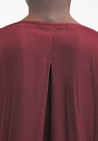 Indumento borgogna che mostra un tessuto liscio e strutturato, con una fessura sul retro e cuciture precise lungo il collo e la cucitura della spalla.